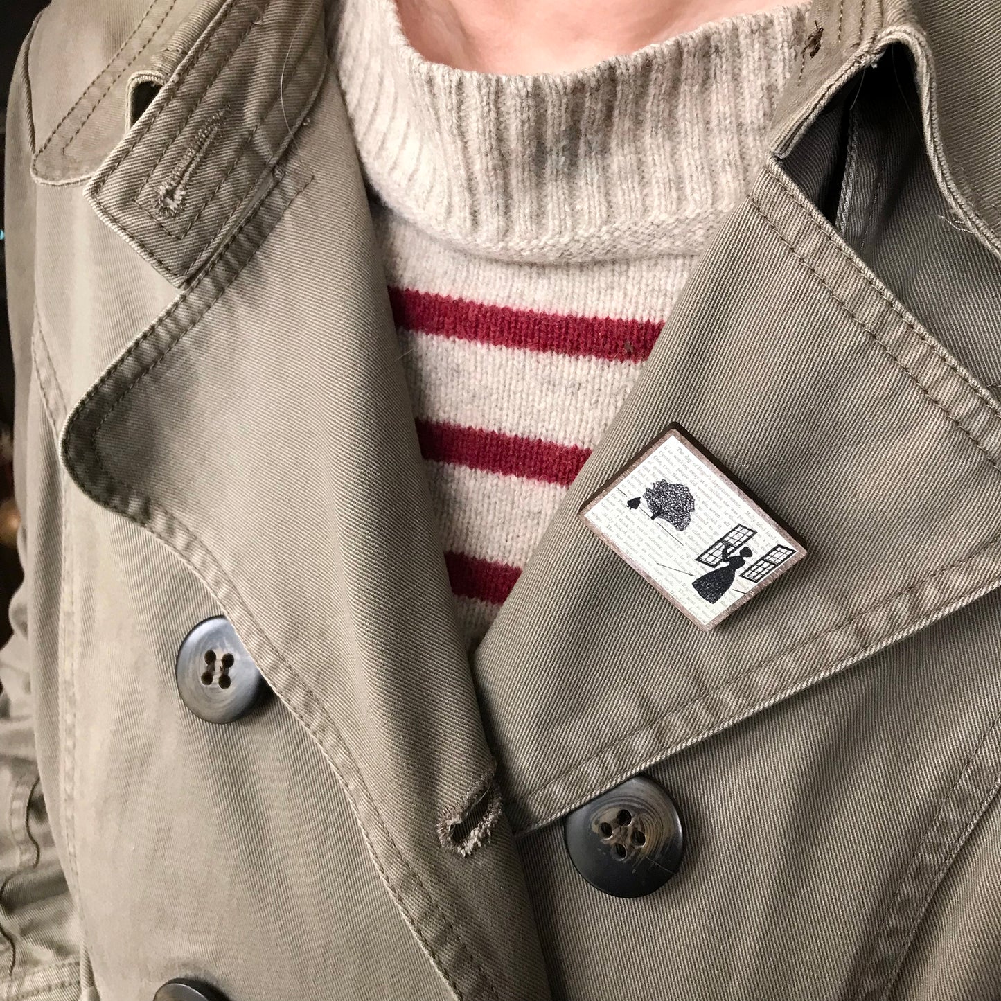 A laser cut wooden brooch by loadofolbobbins showing a silhouette scene inspired by the novel Wives and Daughters by Elizabeth Gaskell and being worn against a blue top. The scene shows Molly looking out of the window at Roger standing under a large tree. The scene is printed against original text from the novel. The brooch is being worn on the lapel of a jacket.