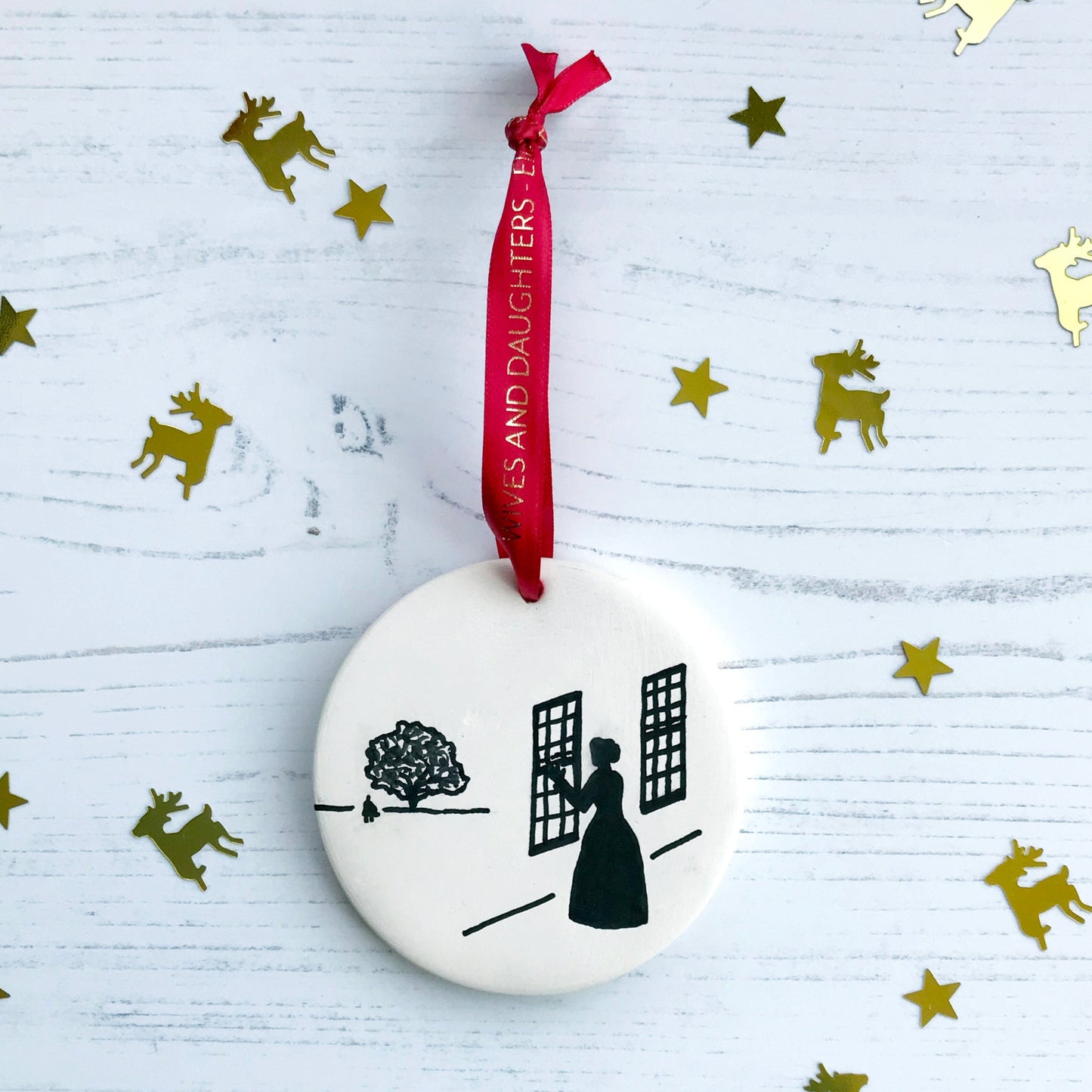 A circular white ceramic hanging decoration showing a hand painted black silhouette scene inspired the novel Wives and Daughters by Elizabeth Gaskell. The scene shows Molly looking out of the window at Roger standing under a large tree. It hangs from a satin red ribbon with gold foil lettering that says Wives and Daughters - Elizabeth Gaskell.