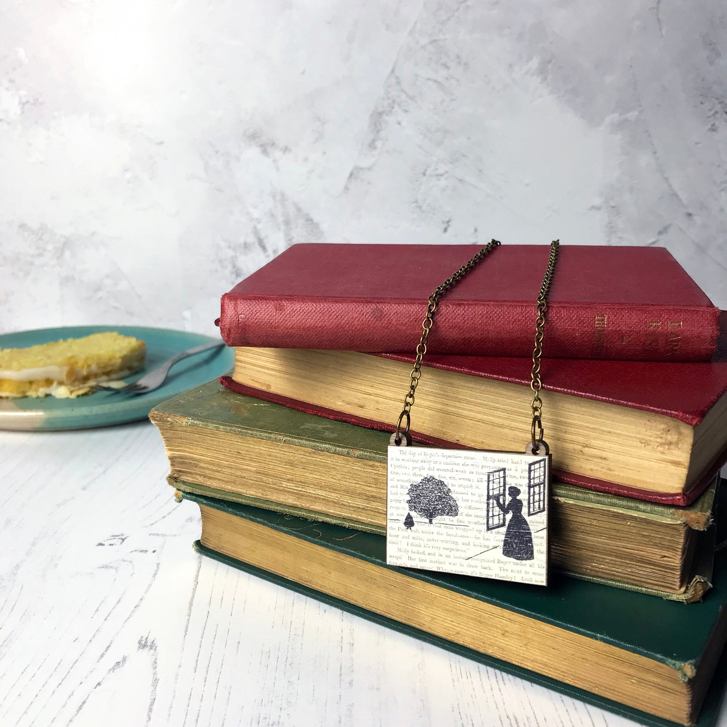 A laser cut wooden necklace by loadofolbobbins showing a silhouette scene inspired by the novel Wives and Daughters by Elizabeth Gaskell. The scene shows Molly looking out of the window at Roger standing under a large tree. The scene is printed against original text from the novel. The necklace is drapped over and dangling on a pile of old books.