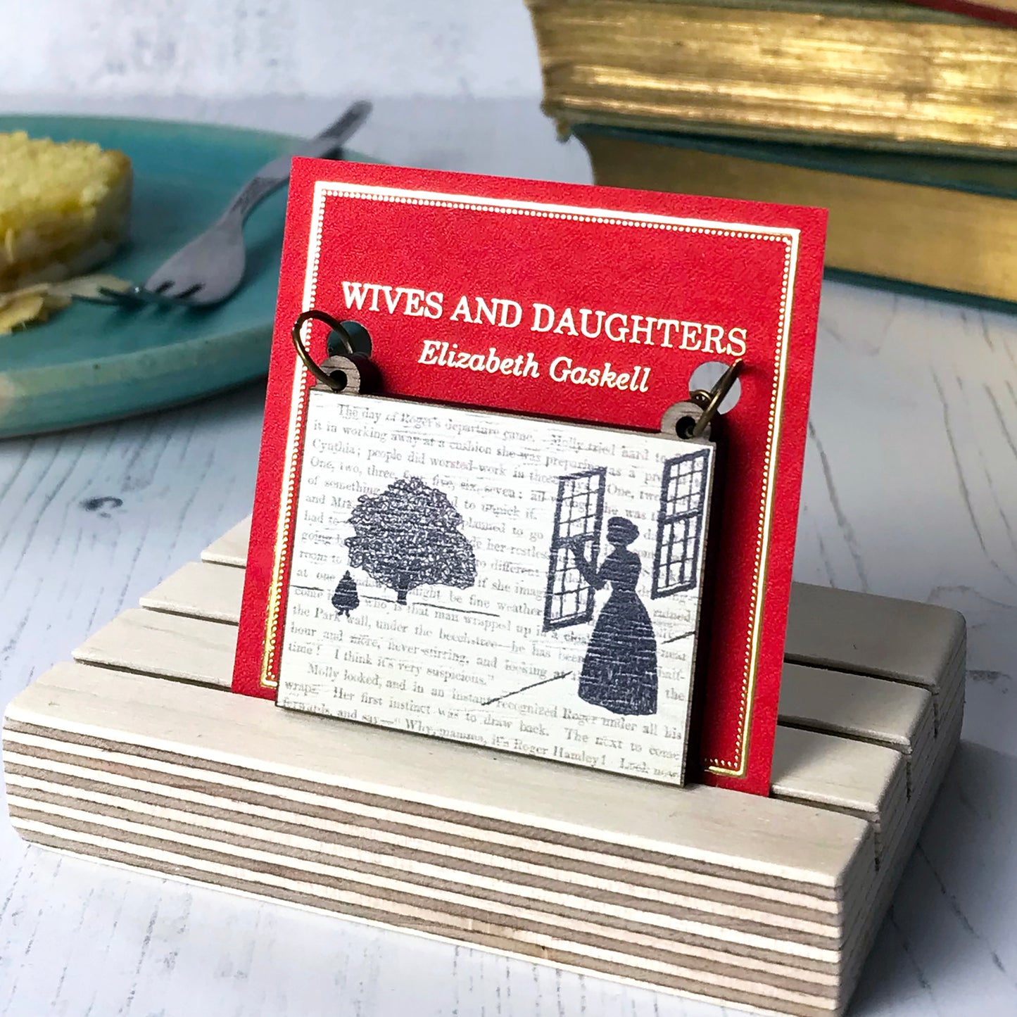 A laser cut wooden necklace by loadofolbobbins showing a silhouette scene inspired by the novel Wives and Daughters by Elizabeth Gaskell. The scene shows Molly looking out of the window at Roger standing under a large tree. The scene is printed against original text from the novel. The necklace is mounted on a red backing card with gold foil lettering detail and made to look like the front of a book.