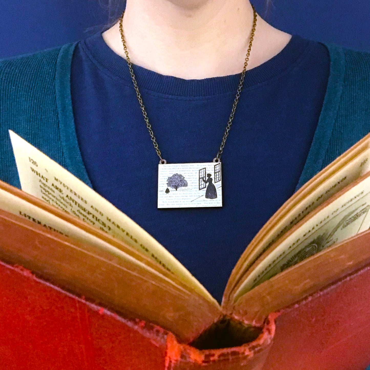 A laser cut wooden necklace by loadofolbobbins showing a silhouette scene inspired by the novel Wives and Daughters by Elizabeth Gaskell and being worn against a blue top. The scene shows Molly looking out of the window at Roger standing under a large tree. The scene is printed against original text from the novel.