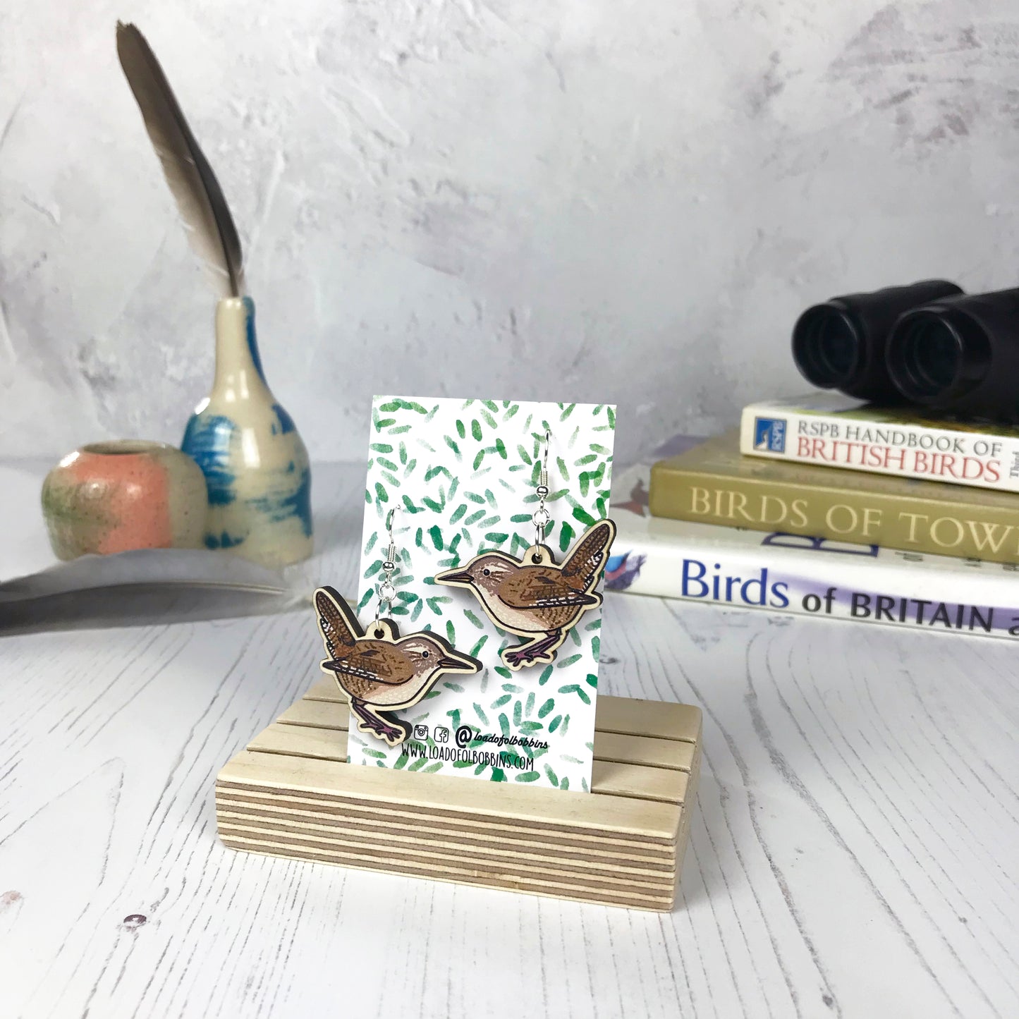 A pair of laser cut wooden dangle earrings showing an illustration of a wren bird by loadofolbobbins. The earrings are mounted on a backing card with a repeat abstract watercolour leaf pattern. They are standing in a wooden stand and in the background you can see a pile of bird watching books and a pair of binoculars.