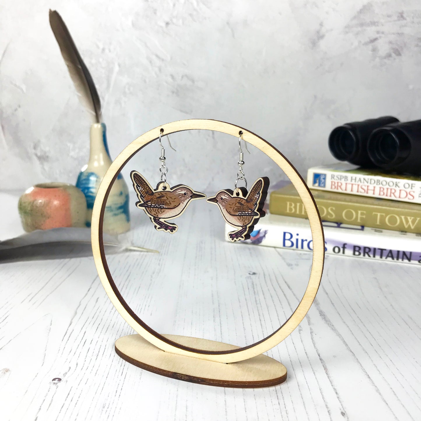 A pair of laser cut wooden dangle earrings showing an illustration of a wren bird by loadofolbobbins. The earrings are hanging from a wooden circular ring display.