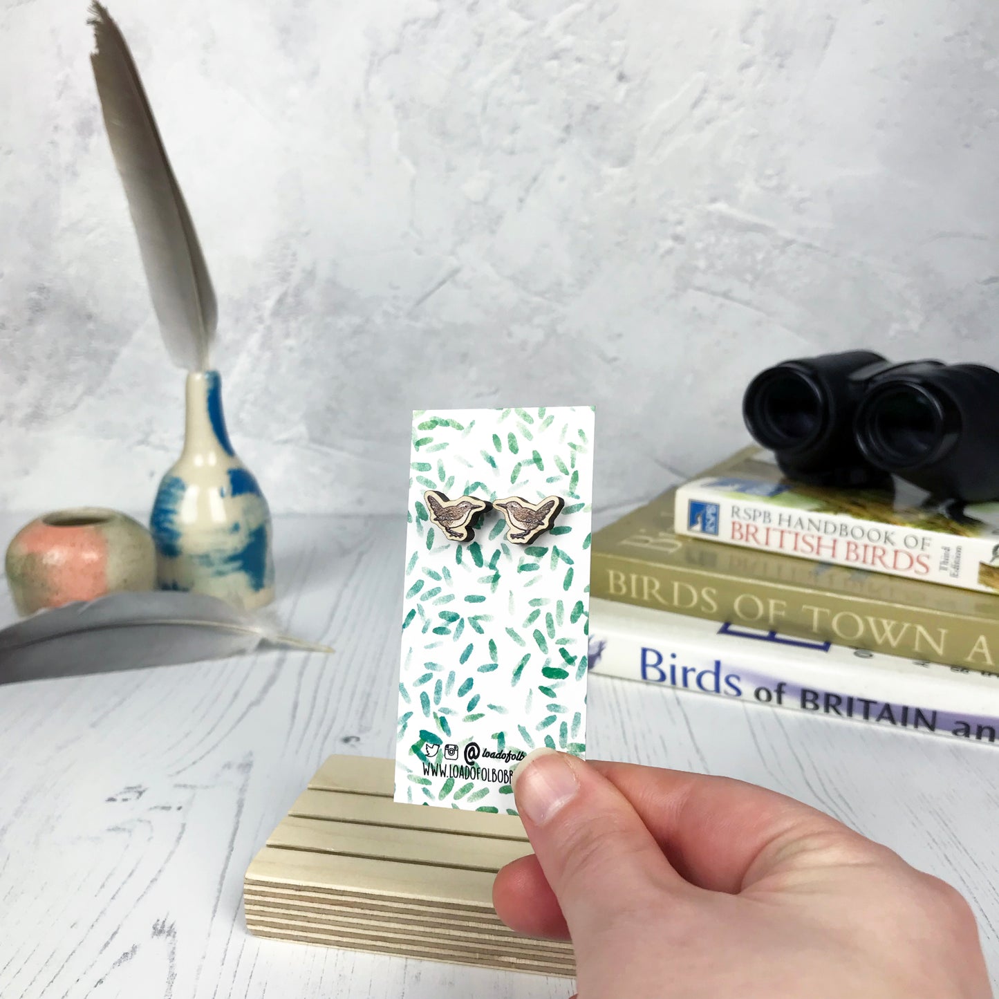 A pair of laser cut wooden earrings showing an illustration of a wren bird by loadofolbobbins. The earrings are mounted on a mini backing card with a repeat abstract watercolour leaf pattern. They are being held by a hand for scale, which shows that each stud earring is slightly smaller than a thumbnail.