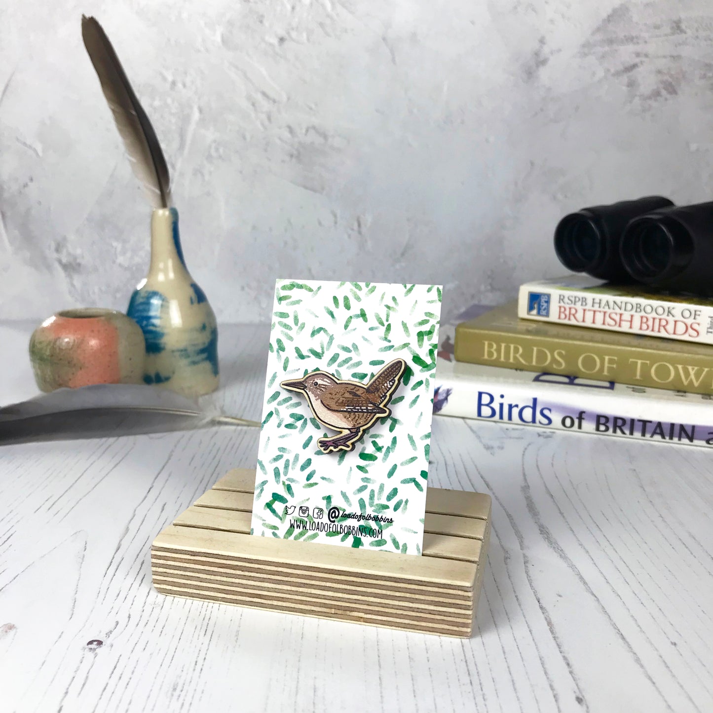 A laser cut wooden brooch showing an illustration of a wren bird by loadofolbobbins. The brooch is mounted on a backing card with a repeat abstract watercolour leaf pattern. They are standing in a wooden stand and in the background you can see a pile of bird watching books and a pair of binoculars.