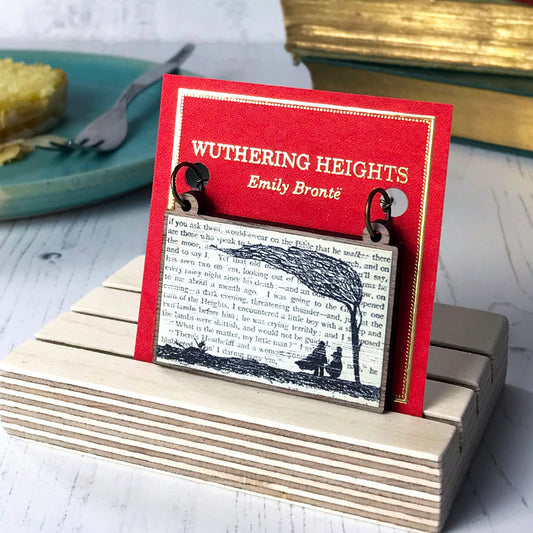 A laser cut wooden necklace by loadofolbobbins showing a silhouette scene inspired by the novel Wuthering Heights by Emily Brontë. The scene shows a silhouette scene of a man and woman under a windswept tree, Heathcliffe and Cathy. The scene is printed against original text from the novel. The necklace is mounted on a red backing card with gold foil lettering detail and made to look like the front of a book.