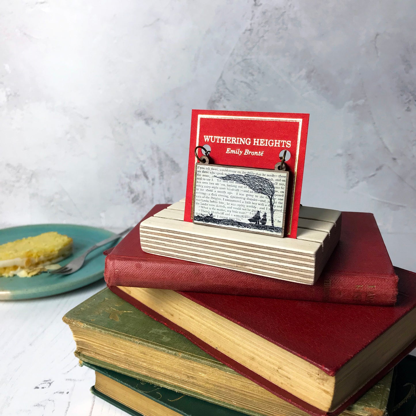 A laser cut wooden necklace by loadofolbobbins showing a silhouette scene inspired by the novel Wuthering Heights by Emily Brontë. The scene shows a silhouette scene of a man and woman under a windswept tree, Heathcliffe and Cathy. The scene is printed against original text from the novel. The necklace is mounted on a red backing card with gold foil lettering detail and made to look like the front of a book and standing on a pile of old books.