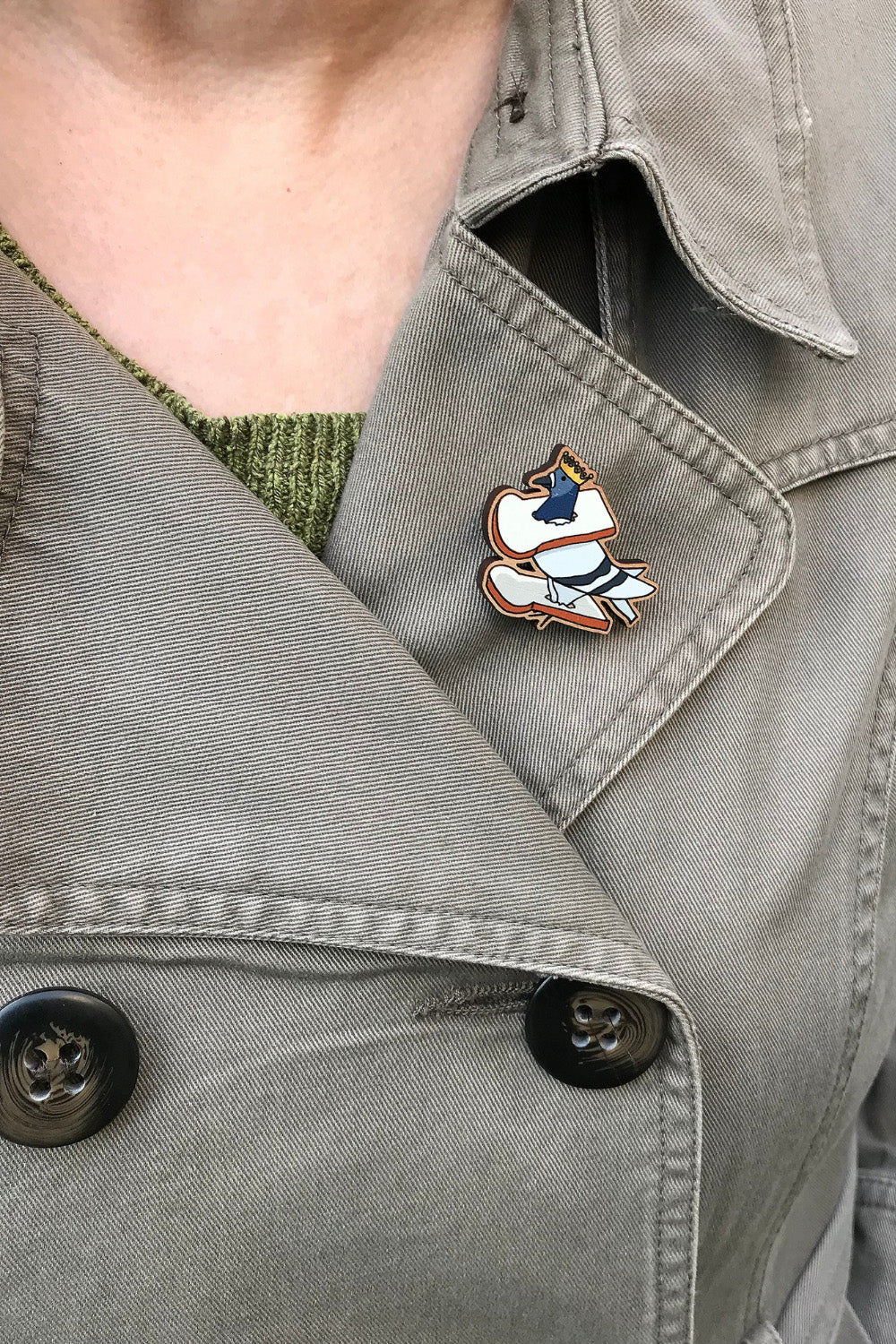 A laser cut wooden lapel pin created from an original illustration by Loadofolbobbins. The pin shows a fun simplified cartoon style illustration of a pigeon with it's feet and head stuck in two slices of bread and also wearing a gold crown. The slice of bread around it's neck is perched around it's shoulders. It's being worn on the lapel of a jacket.