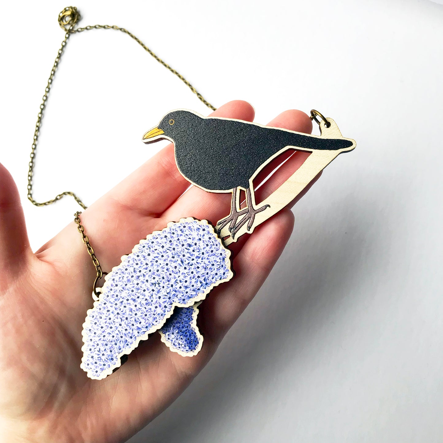 A laser cut wooden charm necklace showing an illustration of a male blackbird perching on a branch with lilac blossoms by loadofolbobbins. The lilac blossom flowers stand proud of the necklace in a 3D relief effect. The wooden necklace is hanging from an antique bronze cable chain and held in a hand for scale, it's about as long as the palm and fingers.
