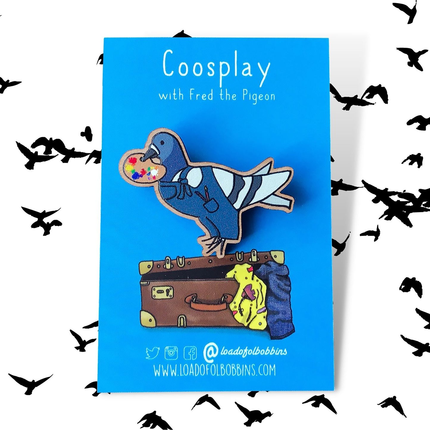 A laser cut wooden lapel pin created from an original illustration by Loadofolbobbins. The pin shows a fun simplified cartoon style illustration of a pigeon dressed as an artist. It's wearing a pair of blue dungarees with paintbrushes in it's mouth and holding a painters pallet full of mixed colours in it's beak. It's is mounted on an illustrated blue backing card which says Coosplay with Fred the pigeon in white text. The pin is made to look like it is standing on a suitcase.