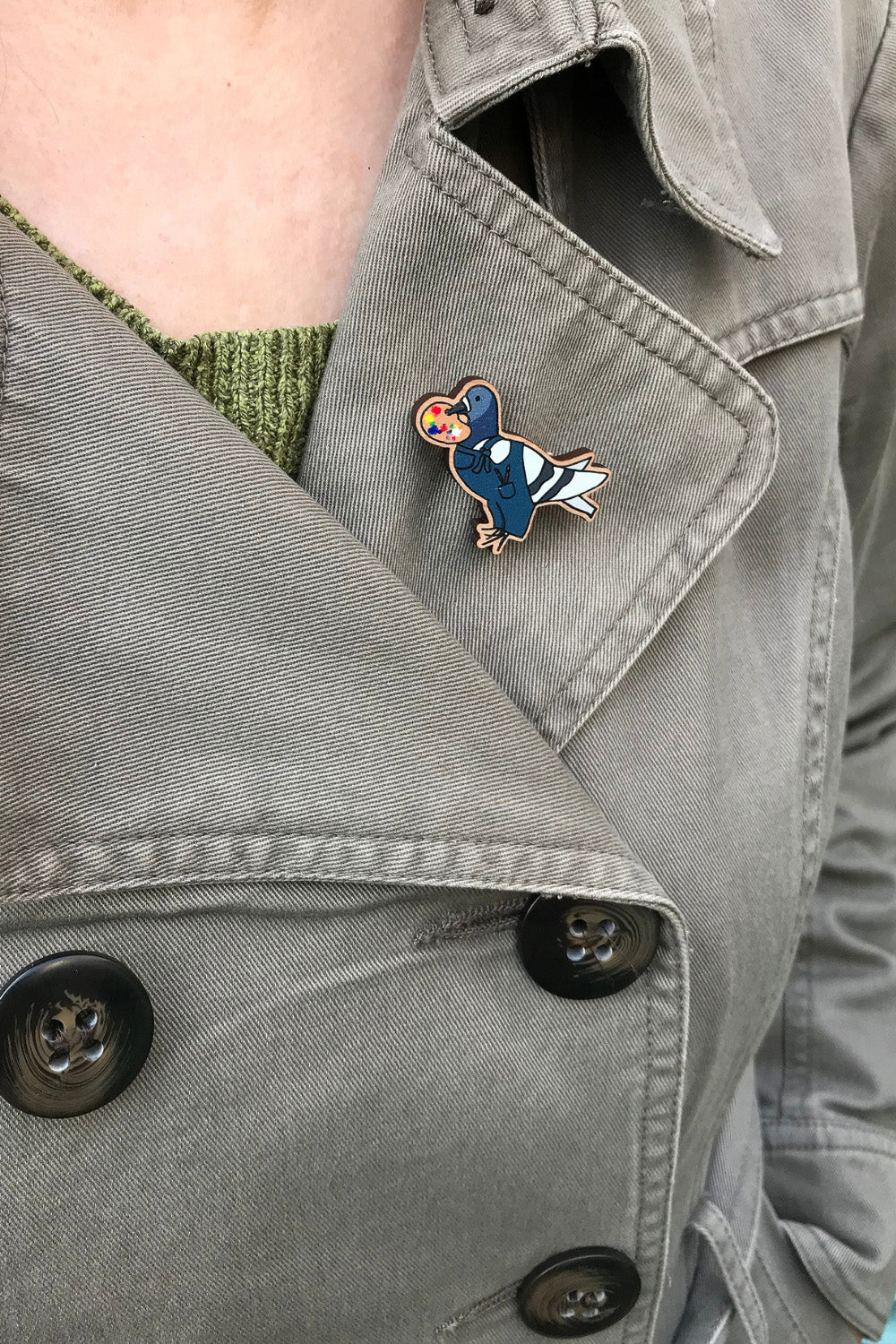 A laser cut wooden lapel pin created from an original illustration by Loadofolbobbins. The pin shows a fun simplified cartoon style illustration of a pigeon dressed as an artist. It's wearing a pair of blue dungarees with paintbrushes in it's mouth and holding a painters pallet full of mixed colours in it's beak. The pin is being worn on the lapel of a jacket.
