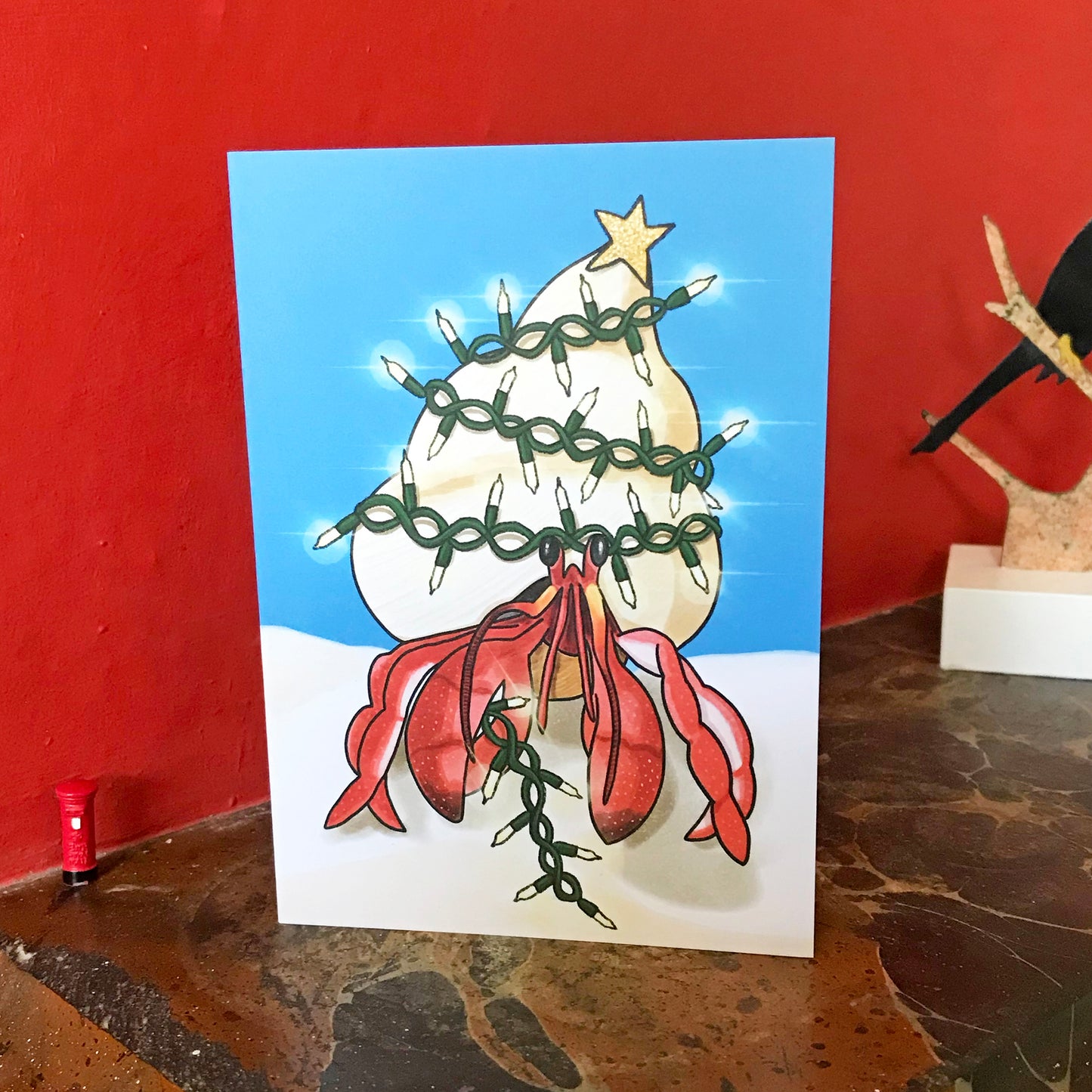 An A6 greetings card showing an illustration by Loadofolbobbins of a hermit crab standing on a sandy beach under vibrant blue sky. Wrapped round their shell is a string of glowing fairy lights that they hold in their right claw. On top of their shell is a glittery gold star, like a Christmas tree. The card is displayed on a mantelpiece.