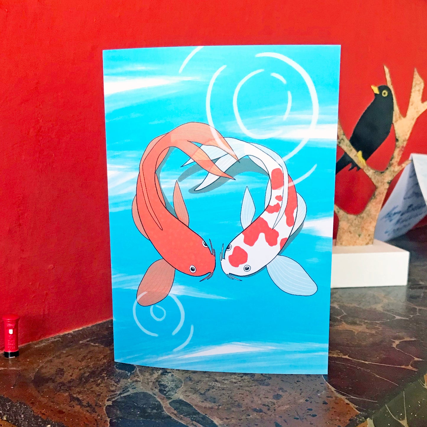 An A6 greetings card with an illustration by Loadofolbobbins of two koi fish facing each other in a pond and forming the shape of a heart. The koi on the left is a vibrant orange and the koi on the right is white with vibrant orange splodges. The background is an energetic light blue line zig zagging down the image with some white showing through. Light white circular marks show ripples on the surface of the water. The card is displayed on a mantelpiece.