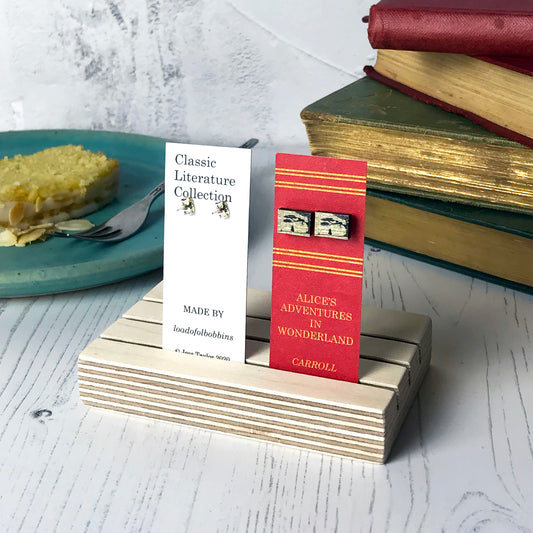 Laser cut wooden stud earrings by loadofolbobbins showing a silhouette scene inspired by the book Alice in Wonderland. It shows Alice coming across the Cheshire Cat who is sitting in the branches of a tree. The scene is printed against original text from the novel. The earrings are mounted on a red backing card with gold lettering detail and made to look like the spine of a book.