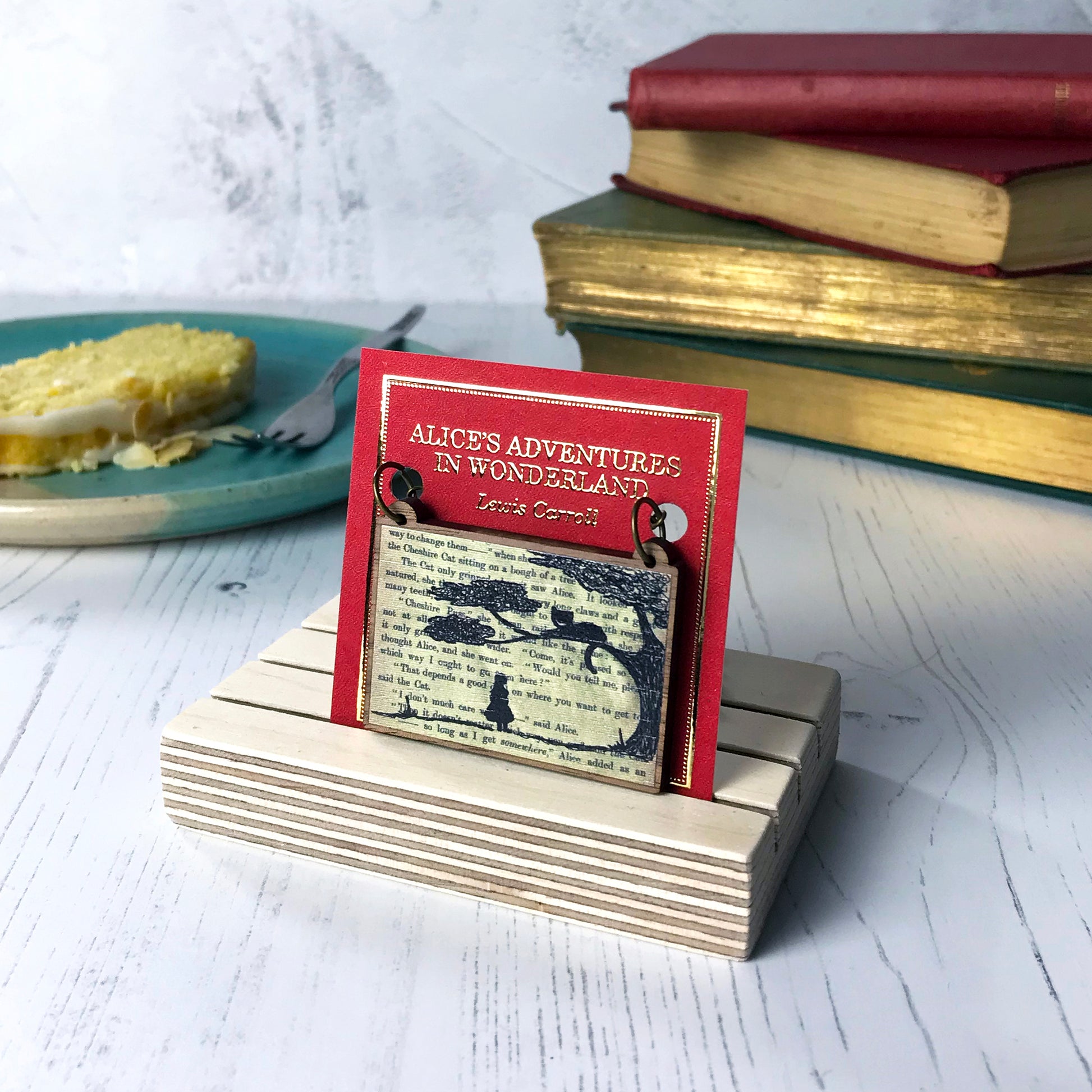 A laser cut wooden necklace by loadofolbobbins showing a silhouette scene of alice looking up at the cheshire cat in a tree against original text from the alice in wonderland novel. The necklace is mounted on a red backing card with gold foil lettering detail saying the name of the novel and writer.