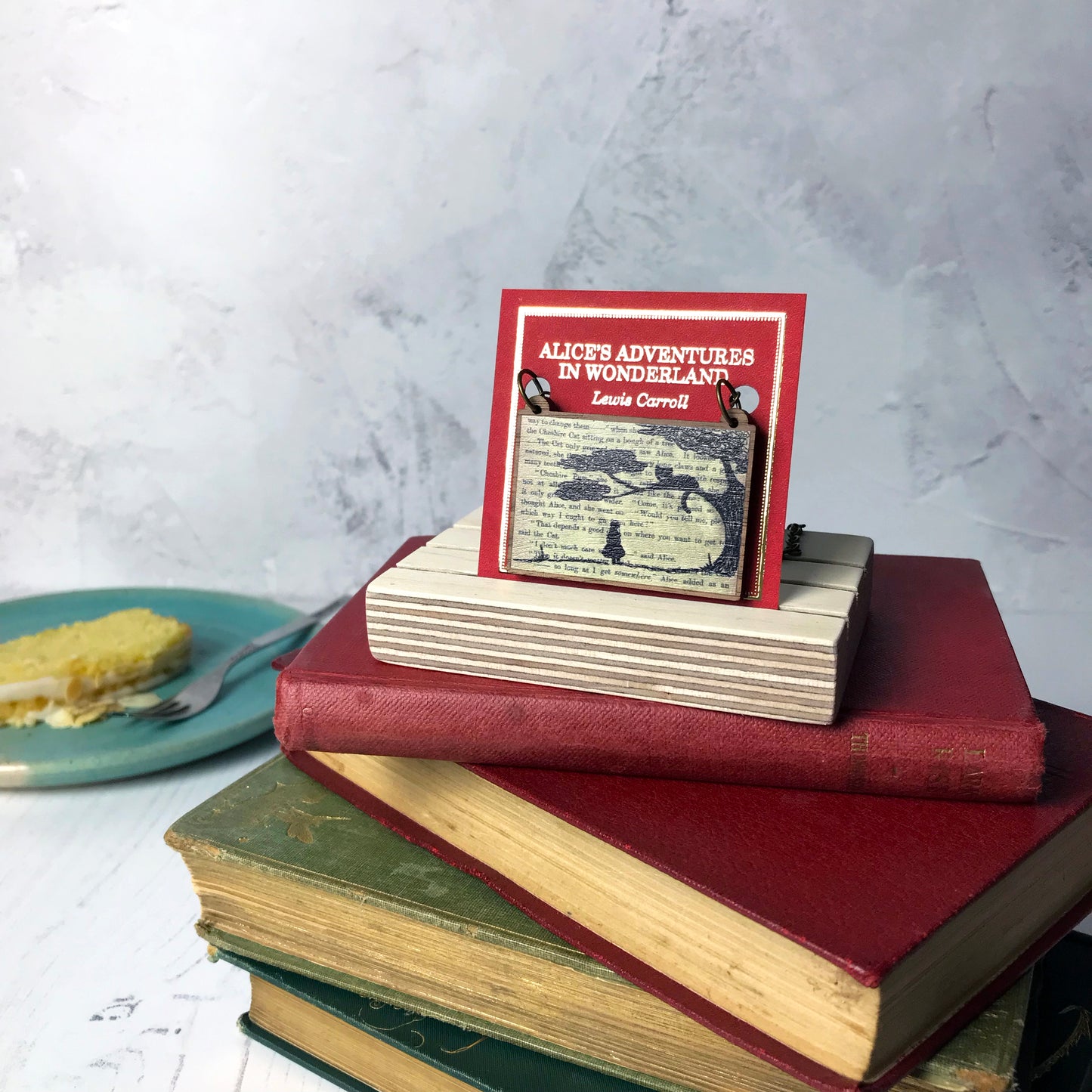 A laser cut wooden necklace by loadofolbobbins showing a silhouette scene of alice looking up at the cheshire cat in a tree against original text from the alice in wonderland novel. The necklace is mounted on a red backing card with gold foil lettering detail saying the name of the novel and writer.