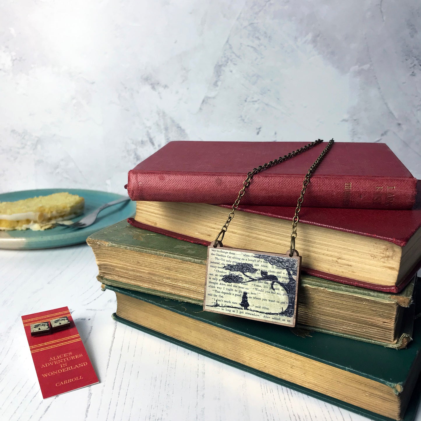 A laser cut wooden necklace and stud earrings by loadofolbobbins showing a silhouette scene of alice looking up at the cheshire cat in a tree against original text from the alice in wonderland novel. The necklace is hung from an antique bronze cable chain and seen dangling over a pile of old books.