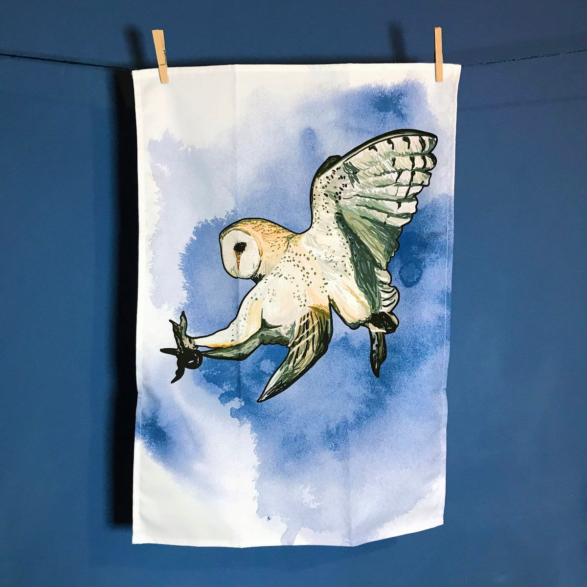 A  photo of  tea towel hanging on a washing line against a dark blue wall. The tea towel shows an illustration of a swooping barn owl against an inky dark blue wash background against white.