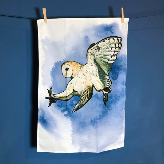 A  photo of  tea towel hanging on a washing line against a dark blue wall. The tea towel shows an illustration of a swooping barn owl against an inky dark blue wash background against white.