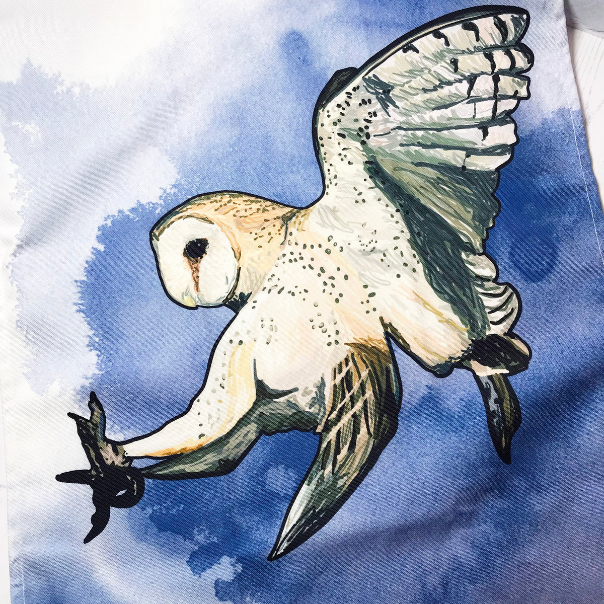 A close up photo of a tea towel showing an illustration of a swooping barn owl against an inky dark blue wash background against white.