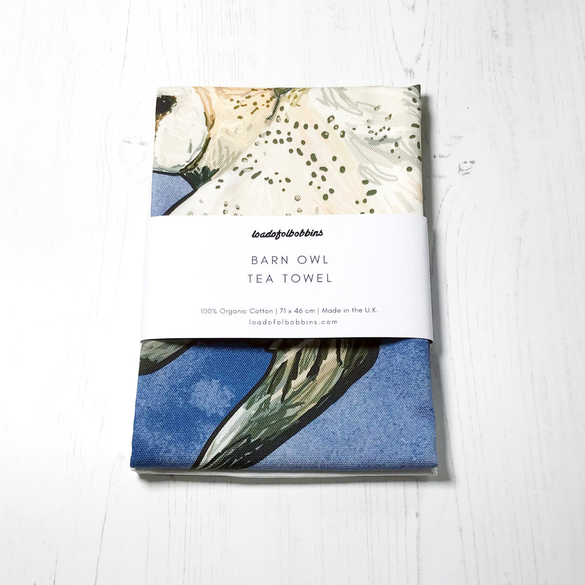 A folded bird themed illustration tea towel by loadofolbobbins. It shows a swooping barn owl against an inky blue wash background.