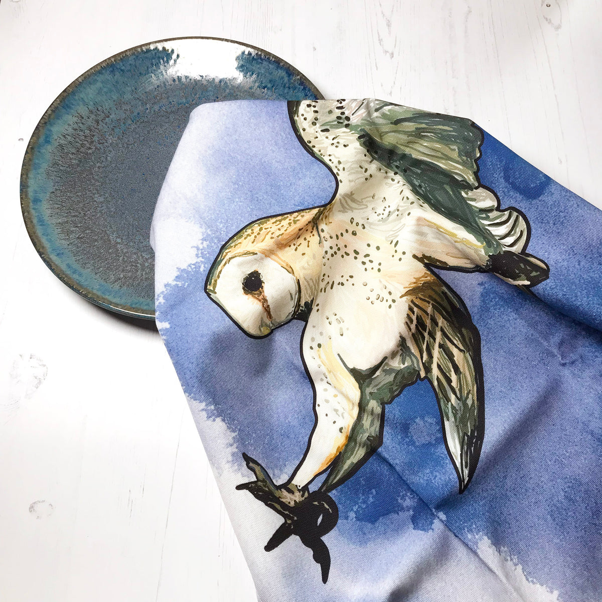 A  photo of a loosely scrunched up tea towel looking like it is drying a ceramic glazed dark turquoise plate. The tea towel shows an illustration of a swooping barn owl against an inky dark blue wash background against white.