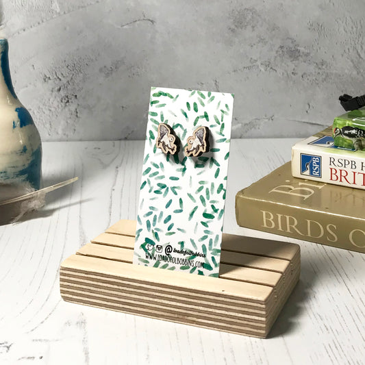 A pair of laser cut wooden earrings showing an illustration of a flying barn owl bird by loadofolbobbins. The earrings are mounted on a mini backing card with a repeat abstract watercolour leaf pattern. They are standing in a wooden stand and in the background you can see a pile of bird watching books and a pair of binoculars.