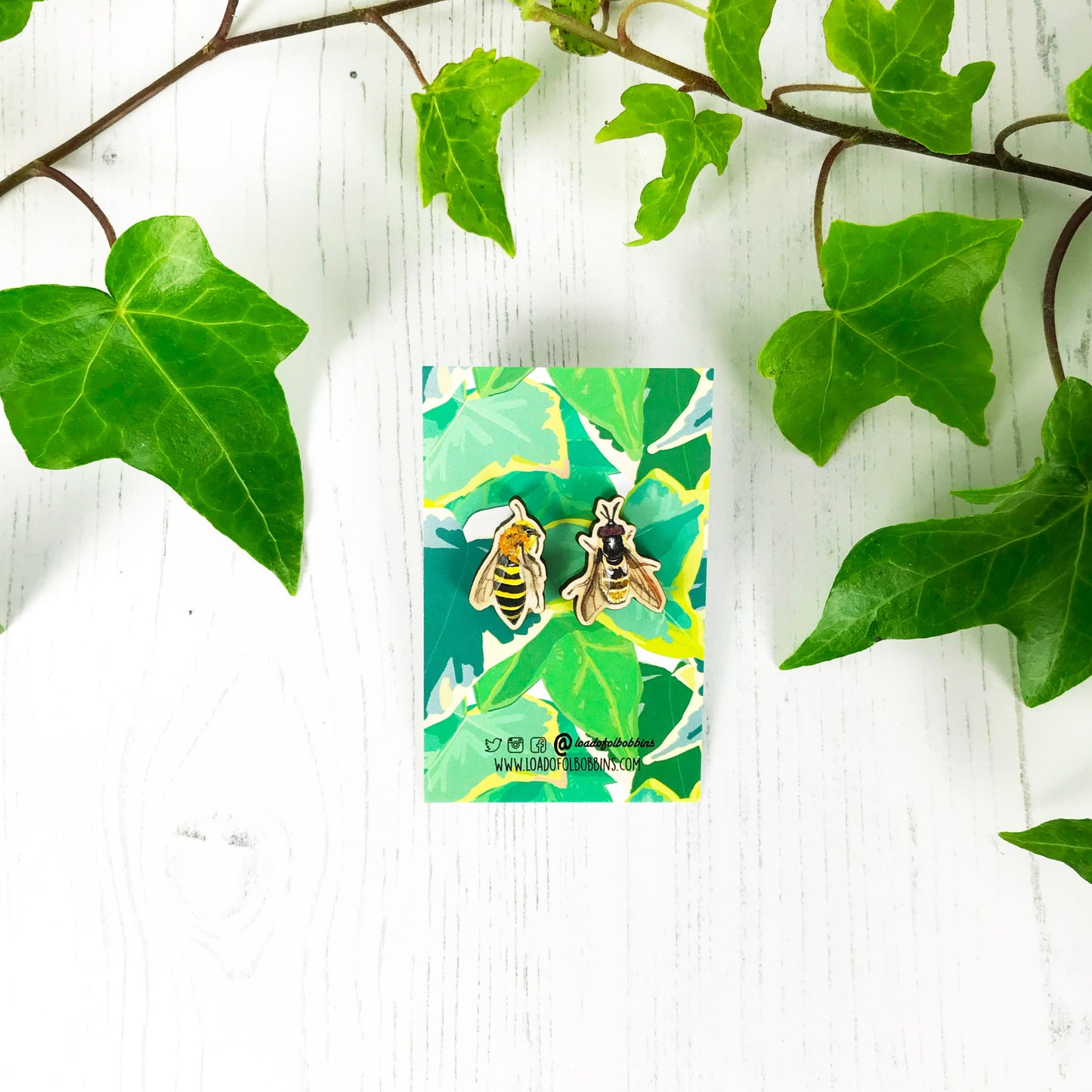 A pair of laser cut mix and match wooden stud earrings showing illustrations by Loadofolbobbins of an ivy mining bee and a golden hoverfly. The earrings are mounted on a backing card with a repeat ivy leaf pattern.