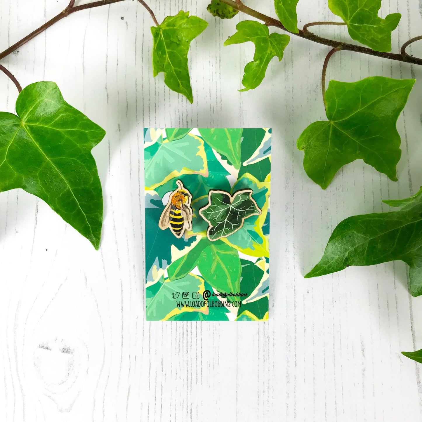 A pair of laser cut mix and match wooden stud earrings showing illustrations by Loadofolbobbins of a dark green ivy leaf and an ivy mining bee. The earrings are mounted on a backing card with a repeat ivy leaf pattern.