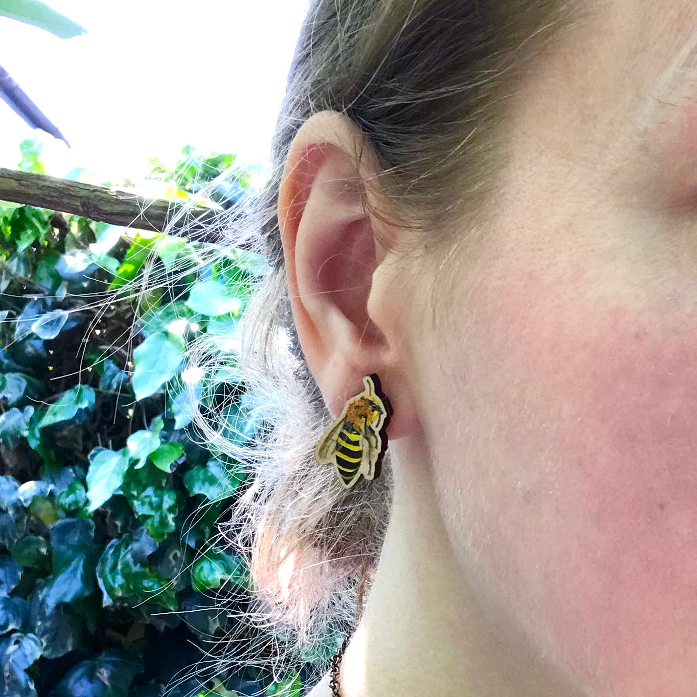 A close up photo of a white woman's ear with blondish brown hair worn up and standing against a wall of ivy. The main focus of the image is her ear which is wearing a small laser cut wooden stud earring of a ivy mining bee by Loadofolbobbins.