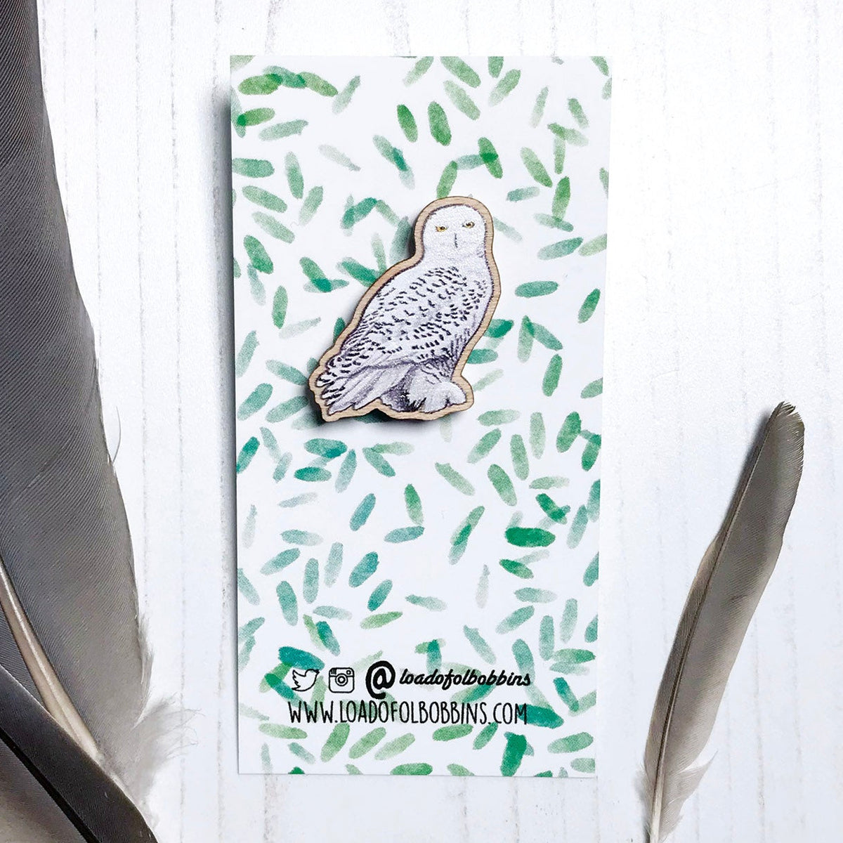 A 25mm mini wooden lapel pin of a snowy owl mounted on an illustrated backing card. The pin is made from an original illustration and is detailed and expressive in style. The bird snow white with brown flecked markings. The backing card has a repeat green watercolour effect pattern that looks like leaves.