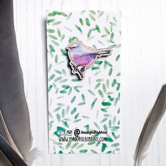 A 26mm mini wooden lapel pin of a white-browed tit warbler mounted on an illustrated backing card. The pin is made from an original illustration and is detailed and expressive in style. The bird is a mix of amazing pinks, purples and blues. The backing card has a repeat green watercolour effect pattern that looks like leaves.