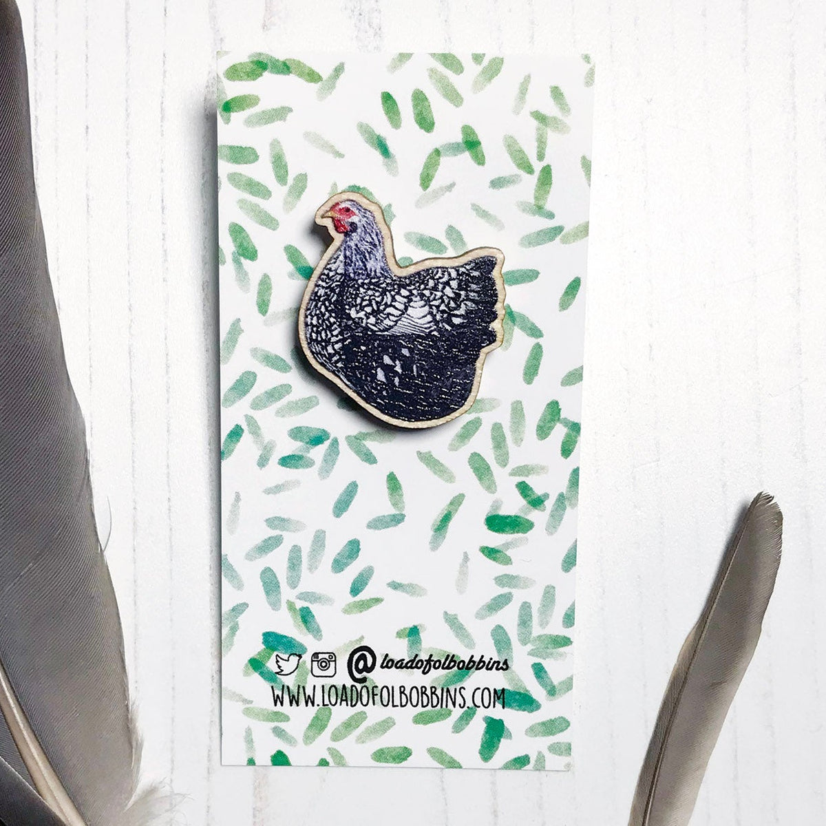 A 26mm mini wooden lapel pin of a Wyandotte chicken mounted on an illustrated backing card. The pin is made from an original illustration and is detailed and expressive in style. The bird is has a lacy black and white pattern. The backing card has a repeat green watercolour effect pattern that looks like leaves.