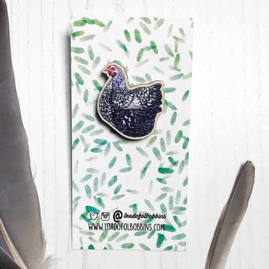 A 26mm mini wooden lapel pin of a Wyandotte chicken mounted on an illustrated backing card. The pin is made from an original illustration and is detailed and expressive in style. The bird is has a lacy black and white pattern. The backing card has a repeat green watercolour effect pattern that looks like leaves.