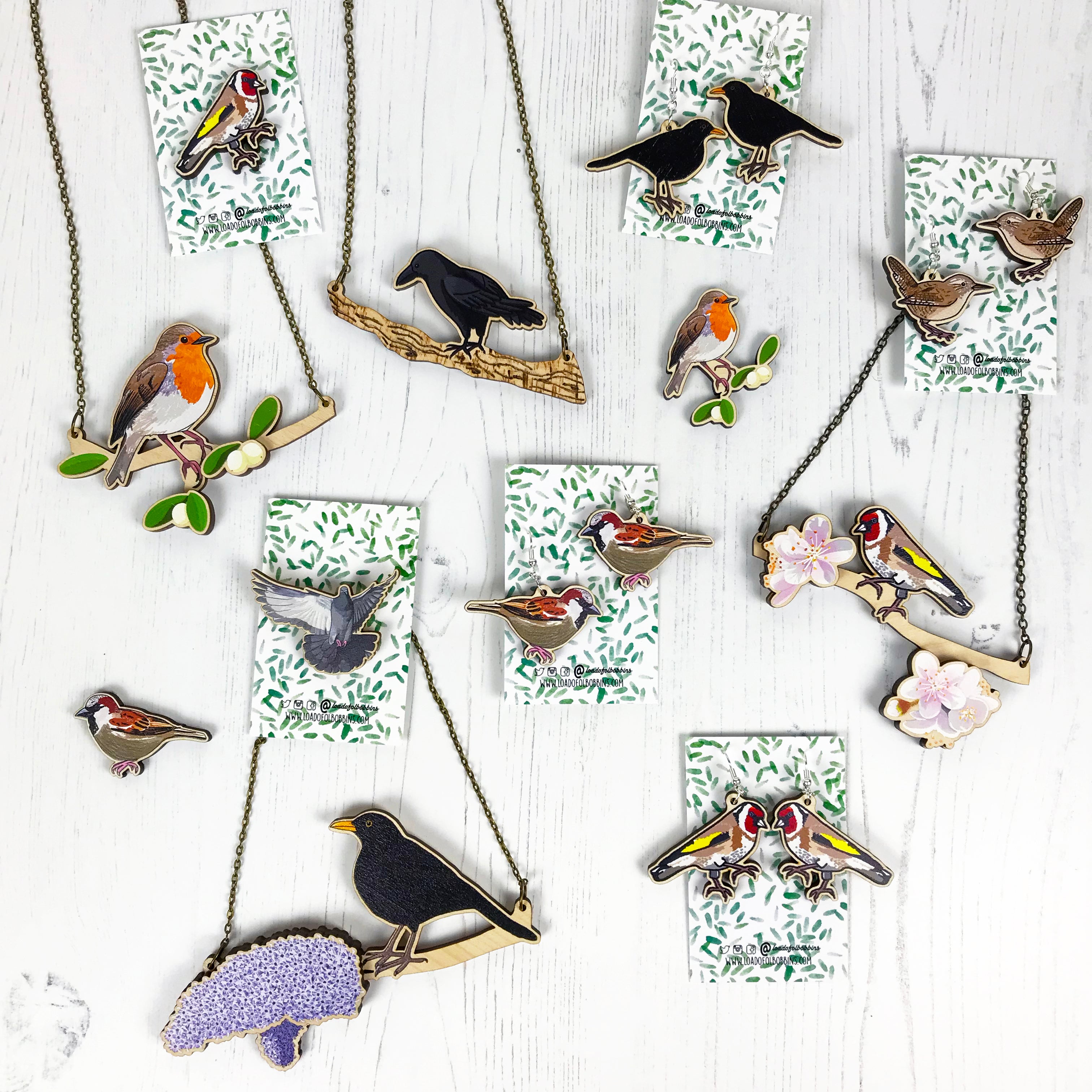 A selection of bird themed laser cut wooden necklaces, earrings and pins from original illustrations by Loadofolbobbins. There's robins, goldfinches, blackbirds, male sparrows, wrens and pigeons. The necklaces have birds perching on branches, goldfinch on cherry blossom with two blooms at either end, male blackbird on a lilac with two big bluey purple blooms, robin on mistletoe with white berries and green leaves, crow is on a branch with pyrography texture and the pigeon is flying with wings out wide.