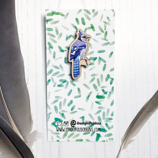 A 33mm mini wooden lapel pin of a Blue jay bird perched on a branch and  mounted on an illustrated backing card. The pin is made from an original illustration and is detailed and expressive in style. The bird is a wonderful array of blues with a small crest on it's head. The backing card has a repeat green watercolour effect pattern that looks like leaves.