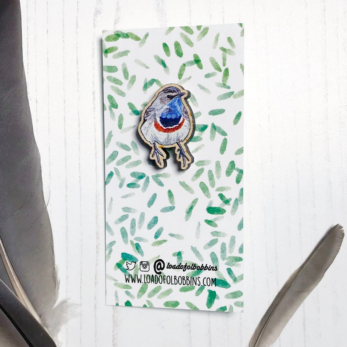A 25mm mini wooden lapel pin of a Bluethroat bird mounted on an illustrated backing card. The pin is made from an original illustration and is detailed and expressive in style. The bird is rather robin like in appearance but with a bright blue throat. The backing card has a repeat green watercolour effect pattern that looks like leaves.