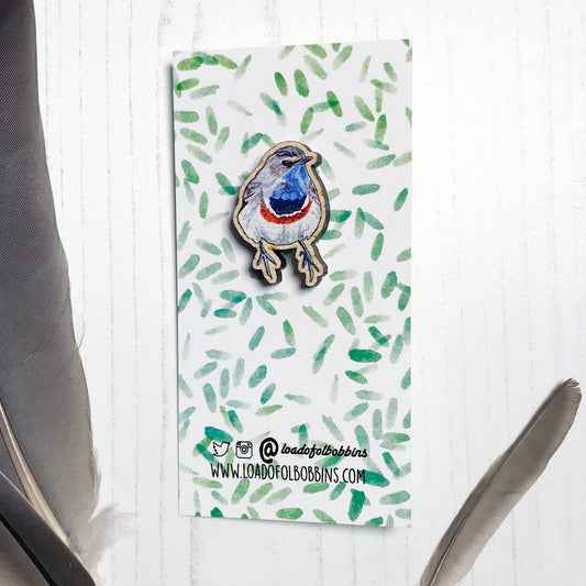 A 25mm mini wooden lapel pin of a Bluethroat bird mounted on an illustrated backing card. The pin is made from an original illustration and is detailed and expressive in style. The bird is rather robin like in appearance but with a bright blue throat. The backing card has a repeat green watercolour effect pattern that looks like leaves.