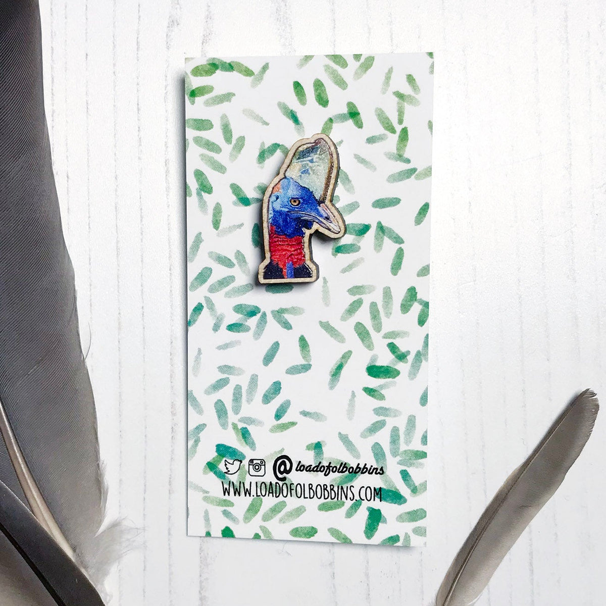 A 25mm mini wooden lapel pin of the head of a Cassowary bird mounted on an illustrated backing card. The pin is made from an original illustration and is detailed and expressive in style. The bird is rather dinosaur like in appearance with a bright blue head and red neck with blue green casque on it's head. The backing card has a repeat green watercolour effect pattern that looks like leaves.