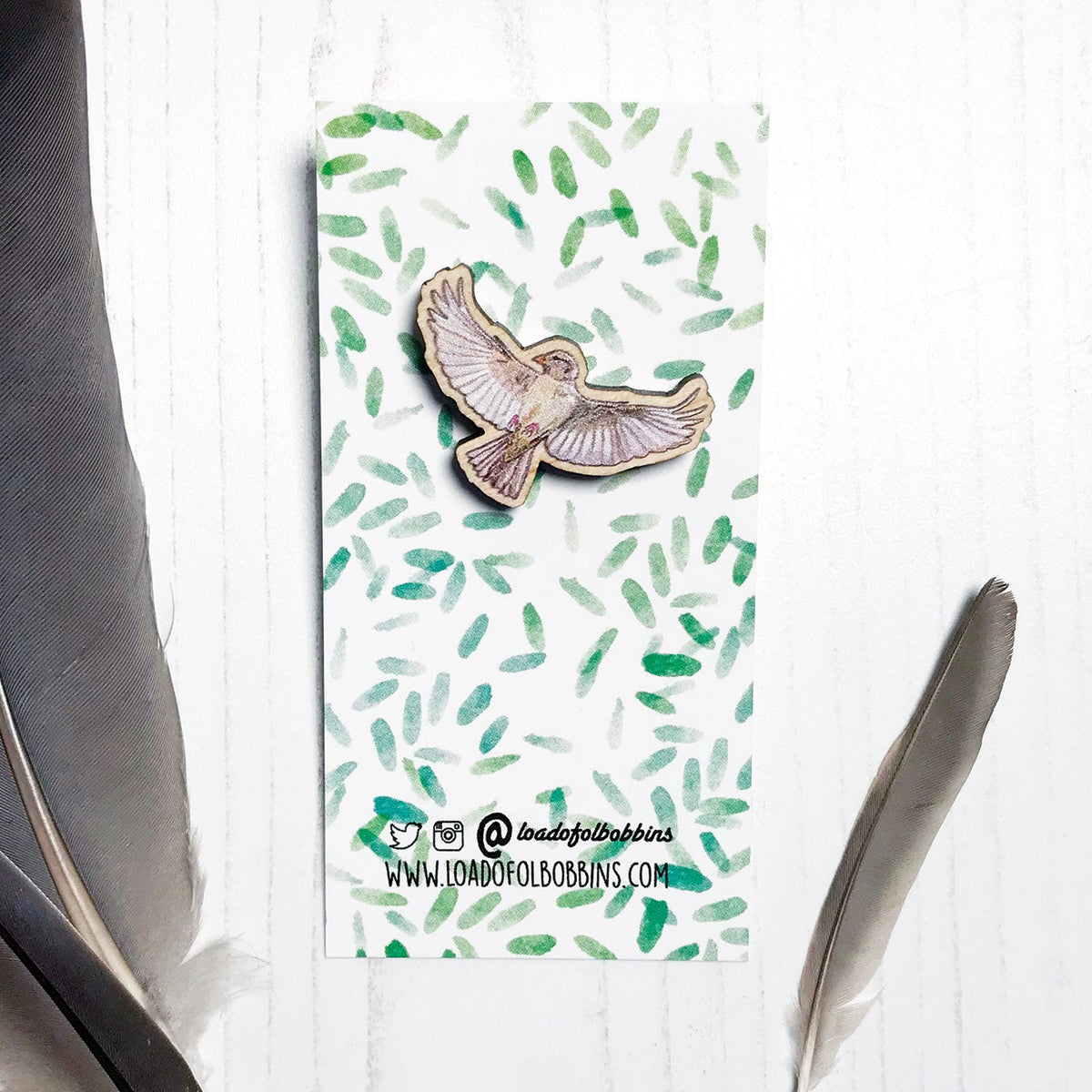 A 30mm mini wooden lapel pin of a flying female sparrow mounted on an illustrated backing card. The pin is made from an original illustration and is detailed and expressive in style. The bird is a gorgeous array of brown hues and has its wings spread wide beside it. The backing card has a repeat green watercolour effect pattern that looks like leaves.