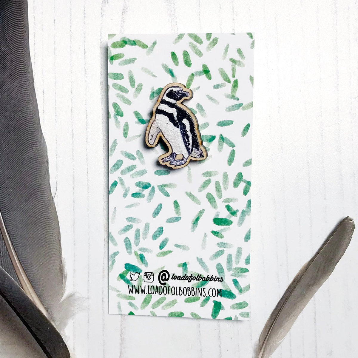A 27mm mini wooden lapel pin of a magellanic penguin mounted on an illustrated backing card. The pin is made from an original illustration and is detailed and expressive in style. The bird has black and white markings. The backing card has a repeat green watercolour effect pattern that looks like leaves.