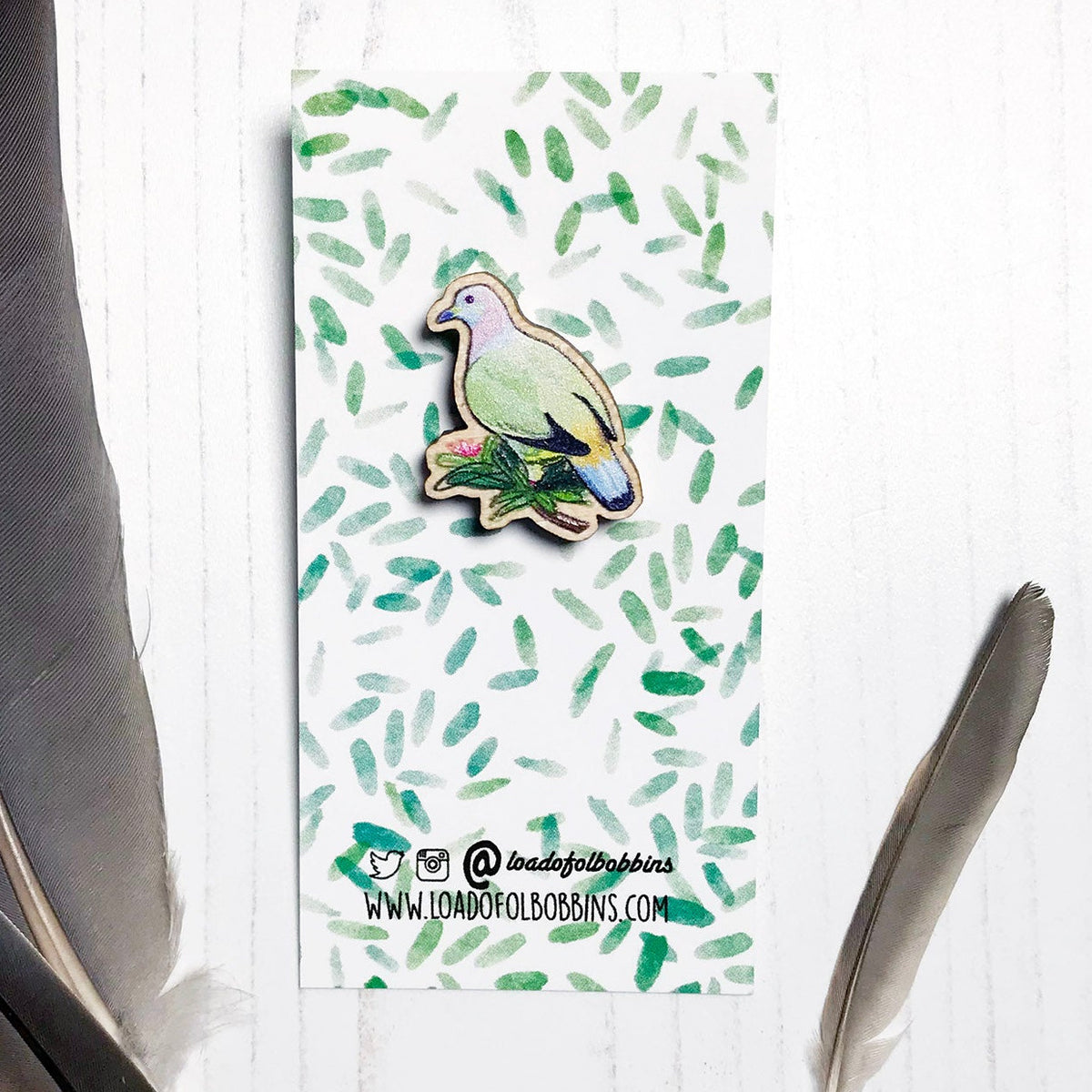 A 27mm mini wooden lapel pin of a pink-necked green pigeon mounted on an illustrated backing card. The pin is made from an original illustration and is detailed and expressive in style. The bird pastel markings in greens, pinks, yellows and blues and is perched on a lush green branch. The backing card has a repeat green watercolour effect pattern that looks like leaves.