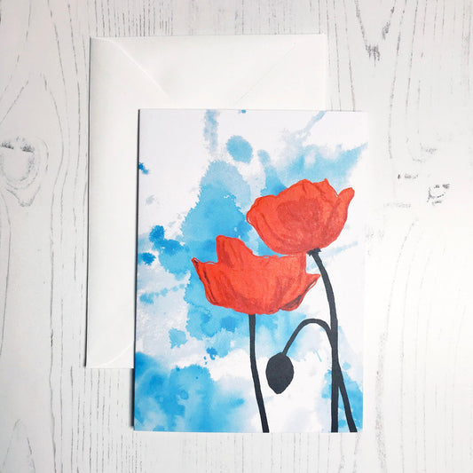 An A6 greetings card showing an illustration of two poppies against an inky blue wash background. The poppies are a vibrant red with the plant stems silhouetted in black.
