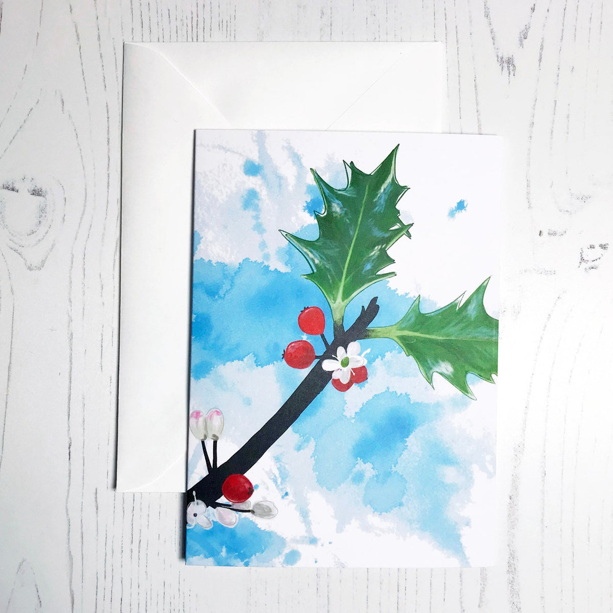 An A6 greetings card showing an illustration of some holly against an inky blue wash background. The holly leaves, berries and flowers are in colour with the plant stems silhouetted in black.