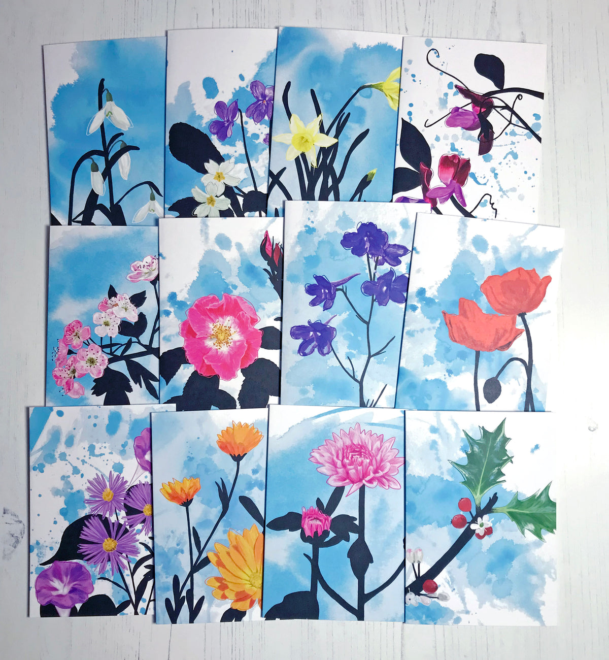 A group photo of twelve different A6 greetings cards showing illustrations representing the different birth flowers for each month against an inky blue wash background. The flowers on each are in colour while the foliage is in black silhouette. From top left they are Snowdrop, Violet and Evening Primrose, Daffordil, Sweet Pea. Hawthorn, Wild Rose, Delphinium, Poppy, Aster and Morning Glory, Marigold, Chrysanthemum and Holly. 