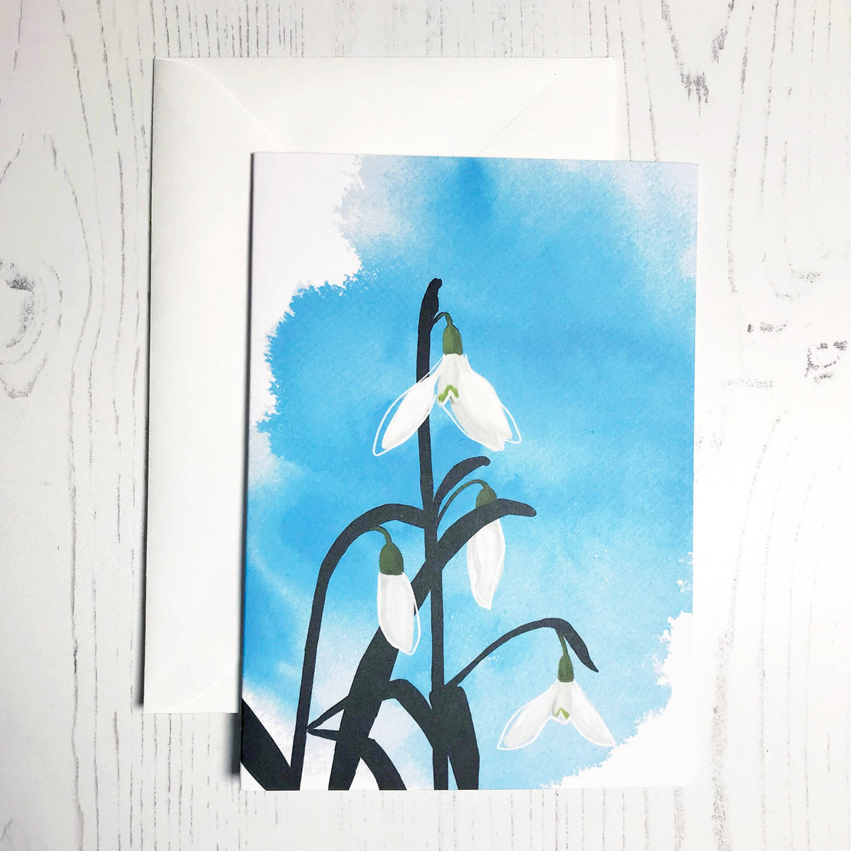 An A6 greetings card showing an illustration of a snowdrop against an inky blue wash background. The snowdrop flowers are in colour with the plant leaves silhouetted in black.