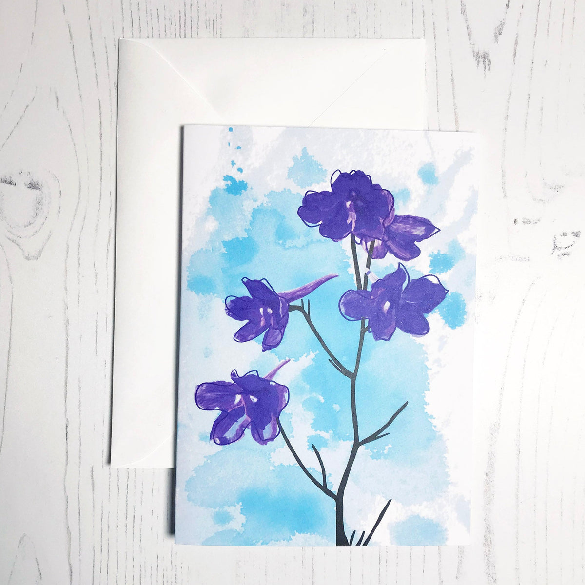An A6 greetings card showing an illustration of delphiniums against an inky blue wash background. The delphinium flowers are in vibrant blue purple colour with the plant stems silhouetted in black.