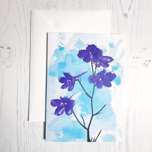 An A6 greetings card showing an illustration of delphiniums against an inky blue wash background. The delphinium flowers are in vibrant blue purple colour with the plant stems silhouetted in black.
