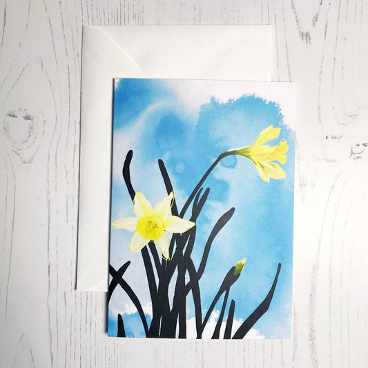 An A6 greetings card showing an illustration of daffodils against an inky blue wash background. The daffodil flowers are in colour with the plant leaves silhouetted in black.