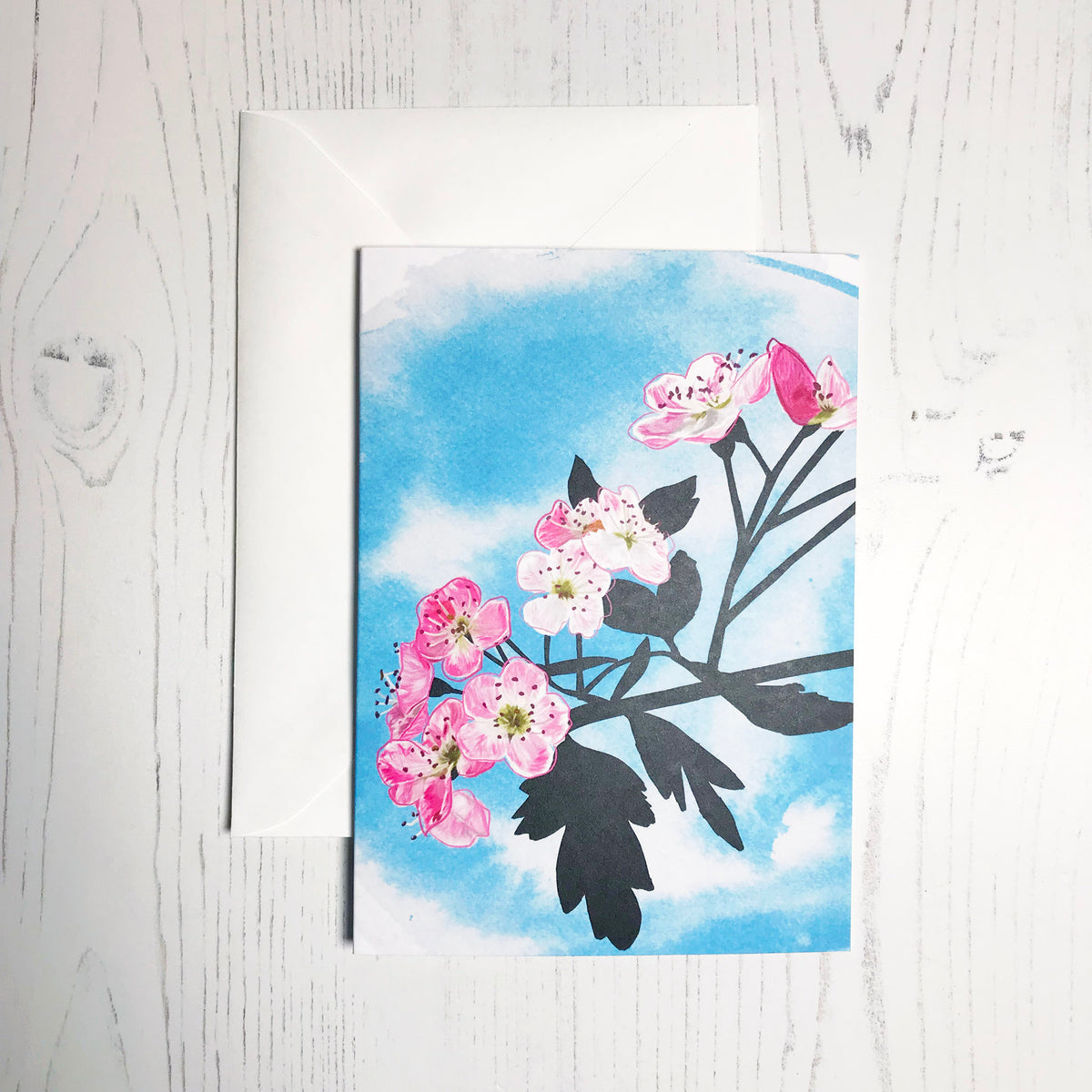 An A6 greetings card showing an illustration of hawthorn blossom against an inky blue wash background. The hawthorn flowers are in colour with the plant leaves and branches silhouetted in black.